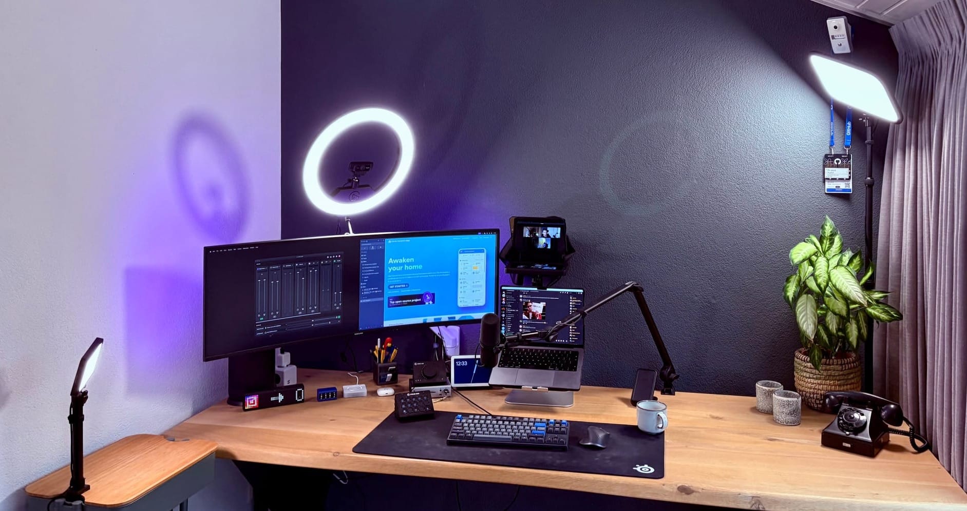 A photo of my office desk. A large wooden desk, with a wide screen and my laptop on it. On the sides are bigger key lights that accommodate for video calling and live streams.