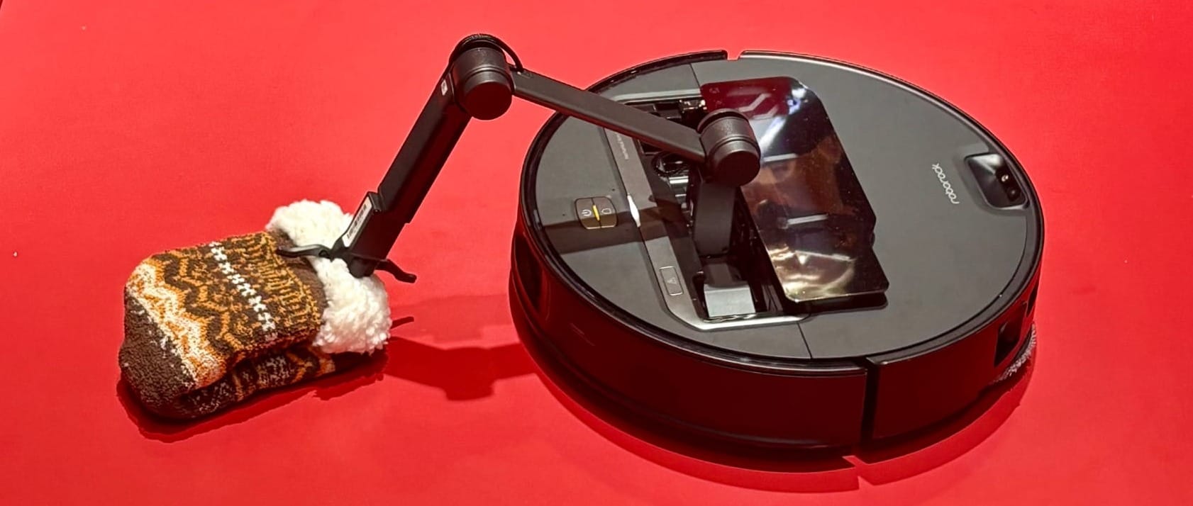 Picture of a robot vacuum, with a small mechanical arm holding a knitted mitten.