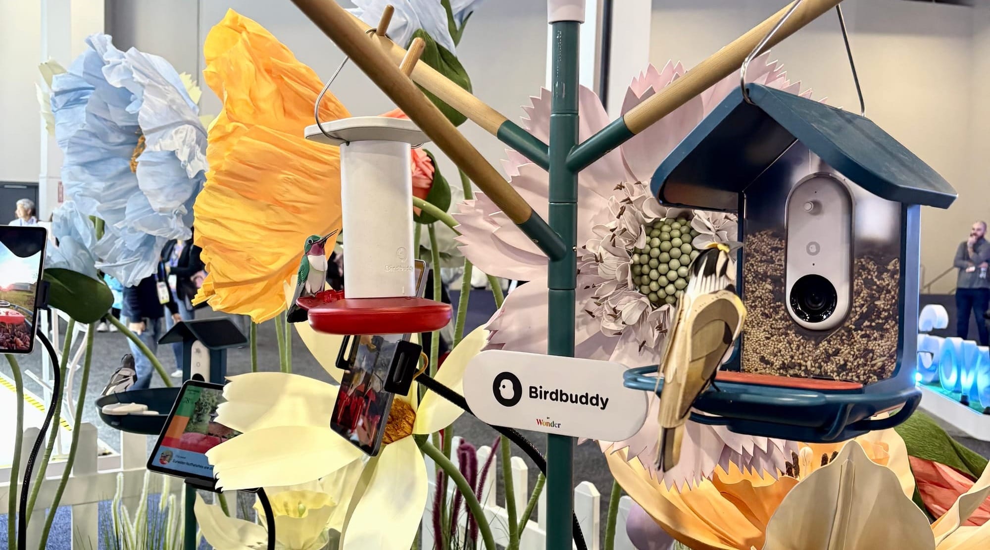 Picture of the Bird Buddy display featuring a camera-equipped feeder with a decorative bird figure, surrounded by oversized, colorful flower props at the CES expo.