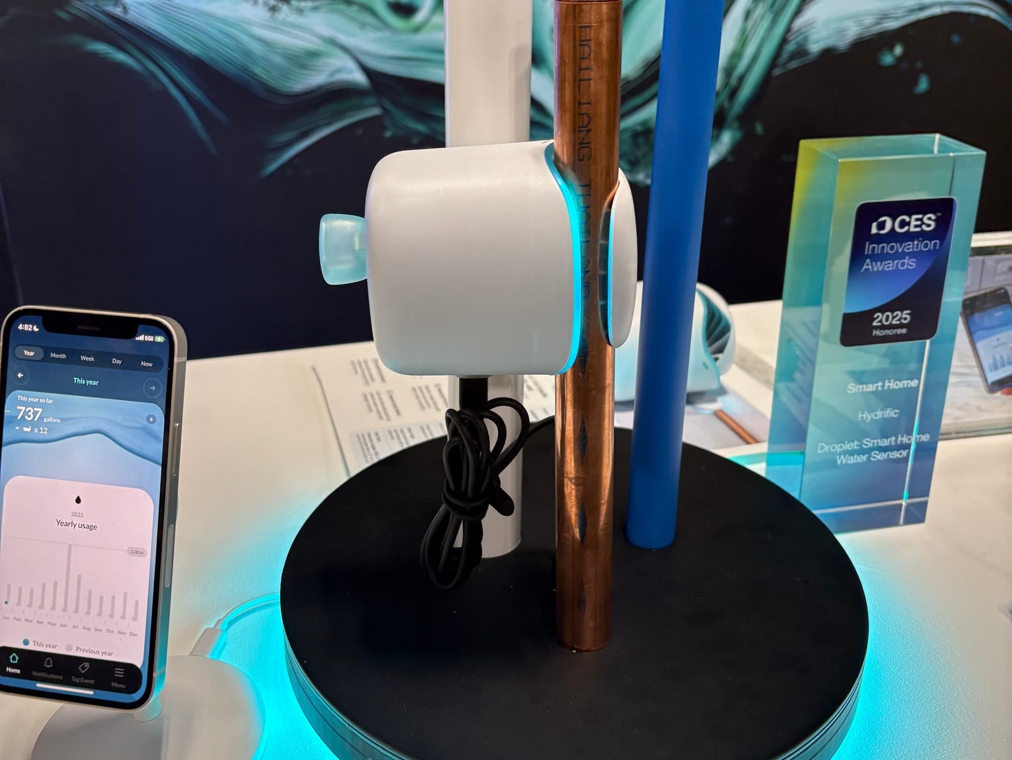 A clamp-on water sensor attached to a copper pipe on a display stand, with a smartphone showing water usage data and a CES Innovation Award in the background.